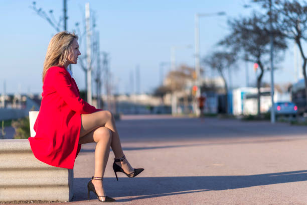 istock Low angle view of a elegant blonde woman in red jacket sitting on a bench outdoors while looking away in sunny day 1176382103
