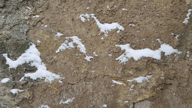 istock Old cracked textured cement wall surface with snow cover on uneven areas. Abstract background in shades of sandy brown color of natural stone. Winter backdrop. Grunge textures. 1176387758