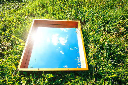 Rectangular mirror lying on the green grass and reflecting blue sky with clouds. Summer, nature trendy concept
