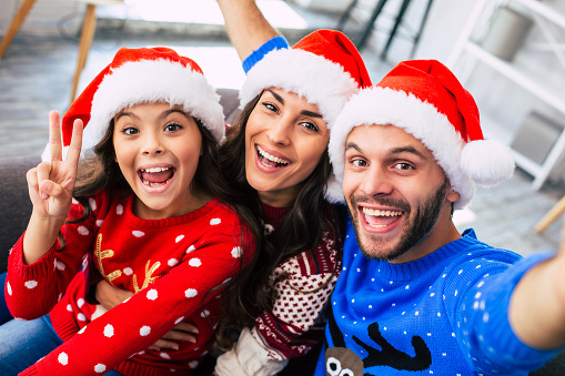 Close-up photo of young mom and dad hugging their little brunette daughter and laughing while taking a selfie in Santa caps at home on a Christmas day