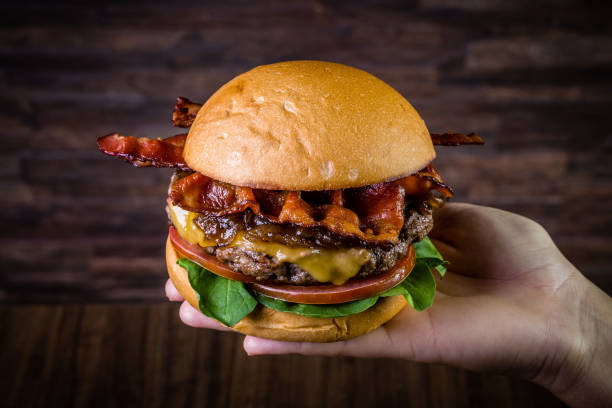 istock Hand holding a craft beef burger with cheese, bacon, caramelized onion and rocket leafs on rustic background 1176402304
