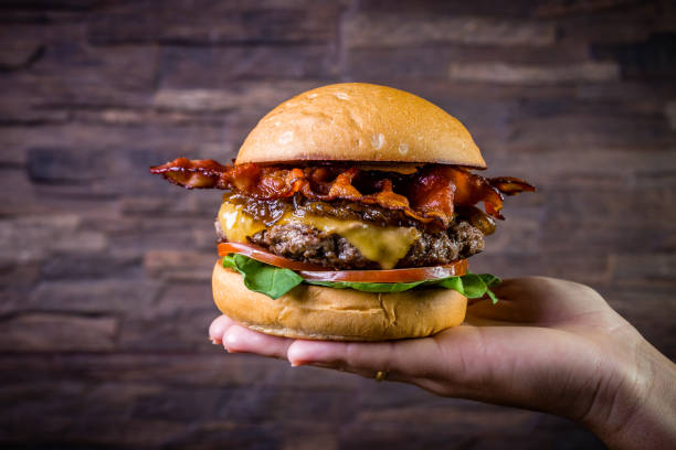 istock Hand holding a craft beef burger with cheese, bacon, caramelized onion and rocket leafs on rustic background 1176403357