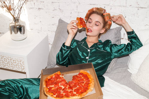 young fashion female eating pizza in bedroom interior young fashionable woman in silk pajamas lays on bed and eat pizza pepperoni from pizza box, direct flash silk photos stock pictures, royalty-free photos & images