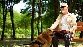 Cheerful blind man holding book and stroking assistance dog, enjoying life