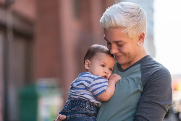 A young adult parents is spending time with their baby. The non-binary gendered adult is carrying the child outside. They are in an urban area near a brick building. The parent is smiling sweetly down at the baby.