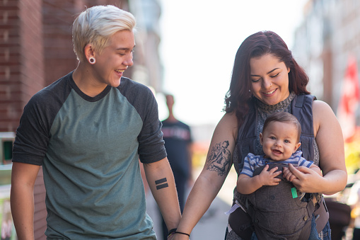 A young adult LGBT couple is spending time together. The partners are walking with their baby in the city. It's a sunny day. The cisgender adult is holding the baby in a carrier. The couple is holding hands and smiling.