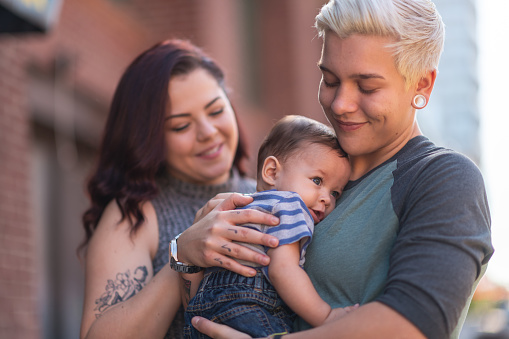A happy LGBT family is spending time together. The young adult partners are with their baby boy outside in a city. They are standing near a brick building. The non-binary parent is holding the child. Both parents are smiling.