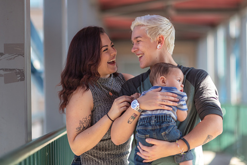 Portrait of a happy LGBT family. The young adult partners are spending time with their baby. The non-binary gendered adult is holding the baby boy. The parents are smiling at each other.
