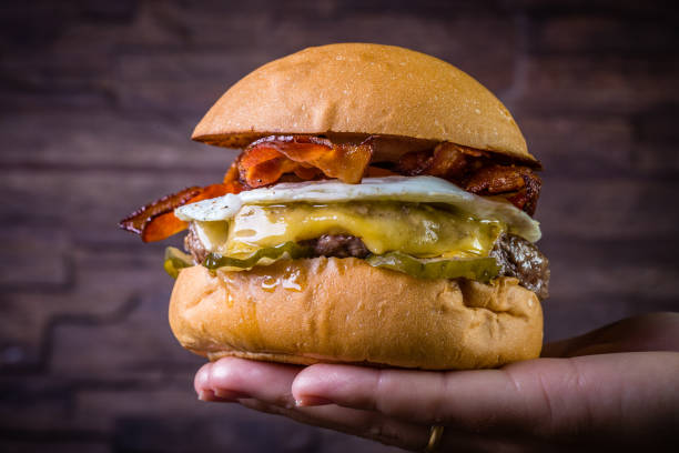istock Hand holding a craft beef burger with cheese, egg, bacon and pickles on wood table and rustic background 1176443915
