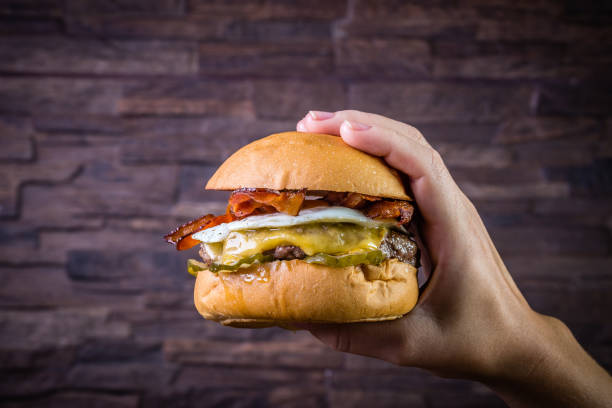 istock Hand holding a craft beef burger with cheese, egg, bacon and pickles on wood table and rustic background 1176446119