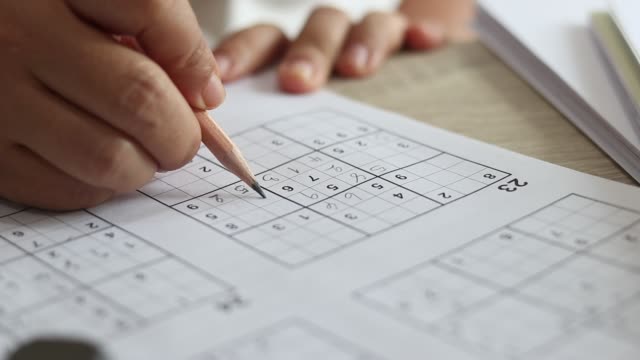 Solve sudoku puzzle with pencil as hobby by senior woman on wooden office desk. Player insert numbers into grid consisting of nine squares subdivided into further nine smaller squares.