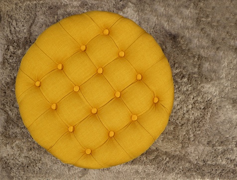 Top view of round fabric yellow pouf with buttons and squares. Gray carpet background .