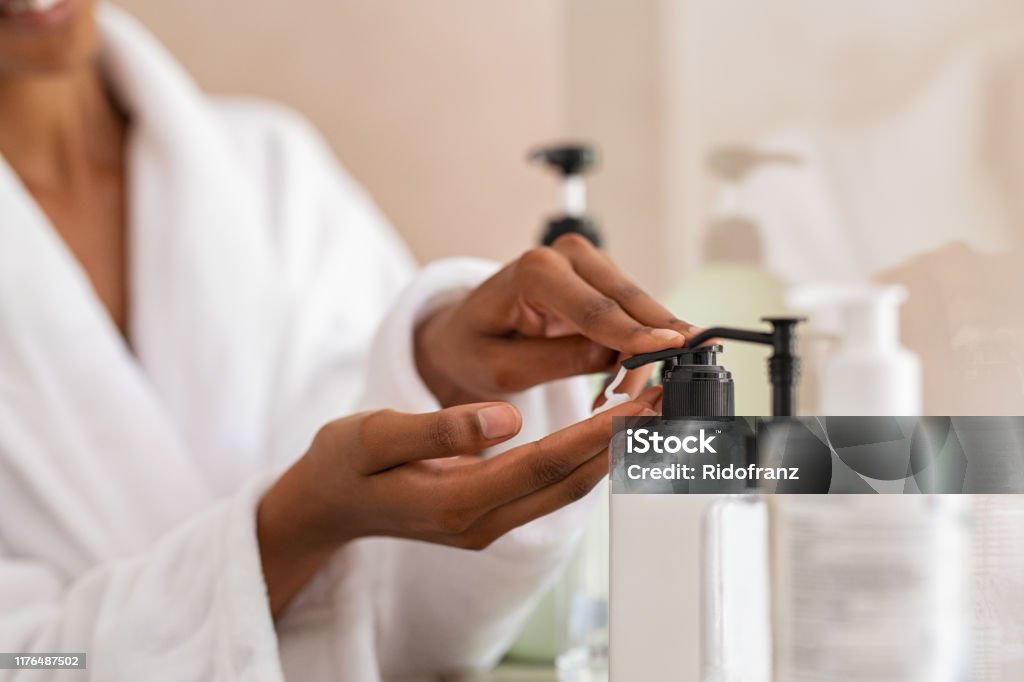 Woman taking body moisturizer from dispenser African woman hands using cosmetic liquid soap in bathroom. Close up of girl black hands in bath robe using body lotion dispenser after shower. Black girl putting pomade on hand from pump. Skin Care Stock Photo