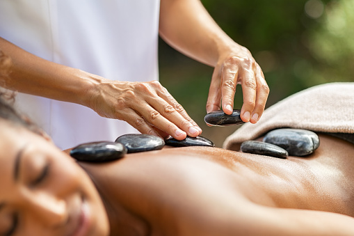 Closeup of masseuse hands placing hot stones on woman back at resort. Masseuse massaging body at spa. Detail of a professional therapist doing lava stones massage on african girl.