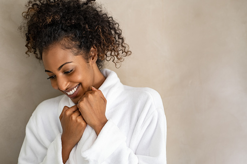 Beautiful young woman in a white bath robe isolated on beige background with copy space. Attractive african girl in bathrobe looking down against light brown wall. Happy african american woman enjoy after body treatment.