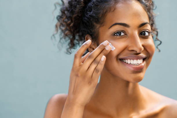 Woman applying moisturizer on face Beautiful african american woman with smooth skin applying moisturizer face cream to her cheek. Beauty young woman taking care of skin. Happy girl applying cosmetic moisturiser treatment isolated on background and looking at camera with copy space. face cream stock pictures, royalty-free photos & images