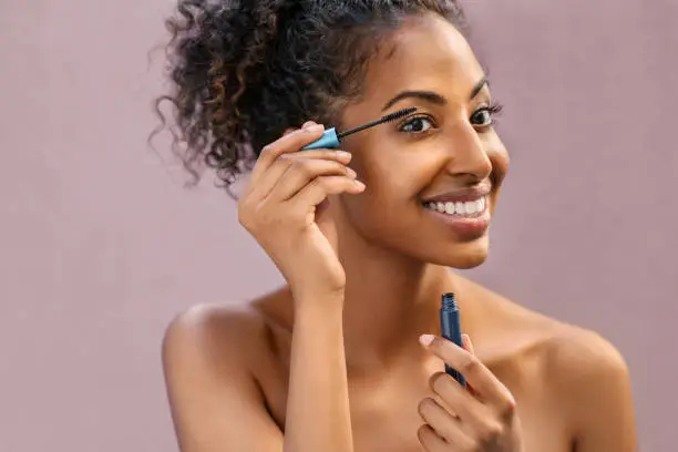 African woman applying black mascara on eyelashes with brush. Young beautiful woman applying makeup on eyes while looking at mirror. Portrait of black beauty girl applying makeup isolated over background with copy space.