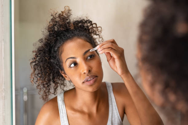 African woman plucking eyebrows African american woman plucking eyebrows with tweezers while standing in front of the mirror. Closeup of young woman in pajamas removing facial hair in bathroom. Girl getting her eyebrows shaped during her morning beauty routine. vanity mirror photos stock pictures, royalty-free photos & images