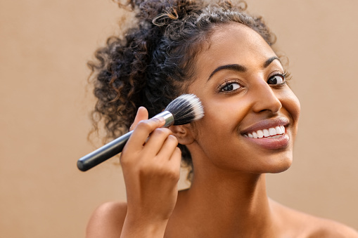 Closeup portrait of african woman applying foundation with makeup brush. Black beauty girl gets blush on the cheek isolated on background. Portrait of attractive young woman looking at camera while using makeup brush for face powder.