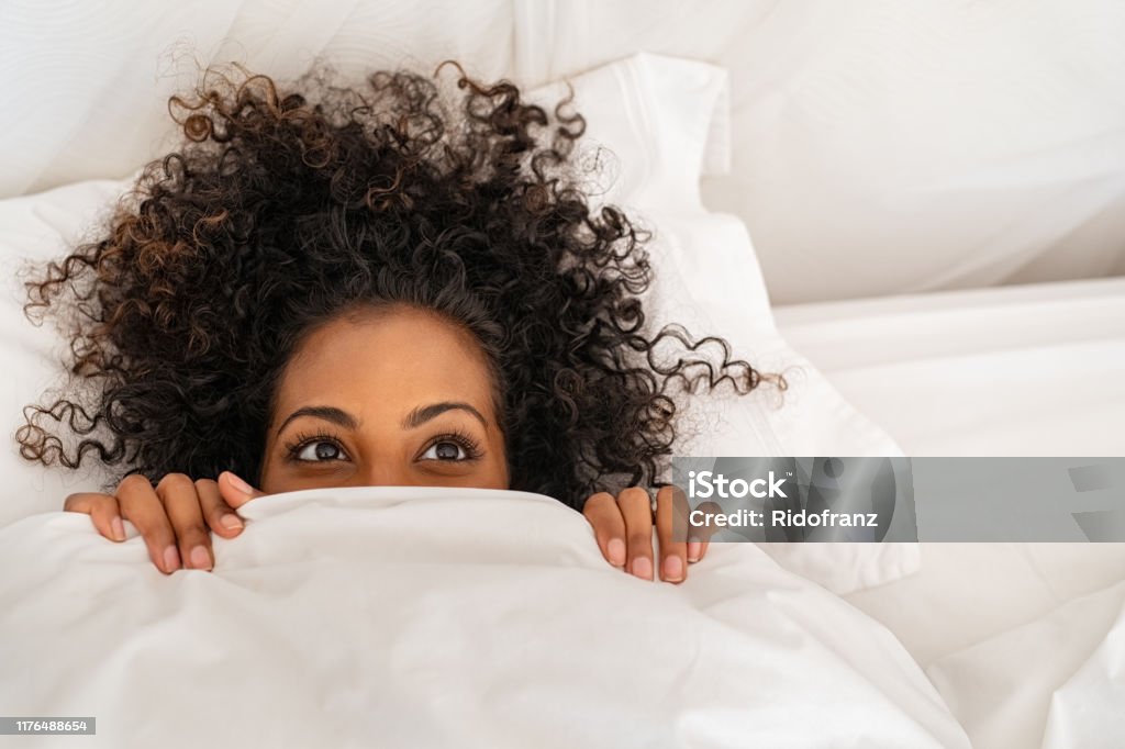 Funny black woman hiding under blanket Funny young woman lying in bed and hiding under sheet while looking up with copy space. Top view of african american girl hiding face under white blanket on bed in the morning. Close up portrait of beautiful woman with curly hair covering face with bed sheet. Bed - Furniture Stock Photo