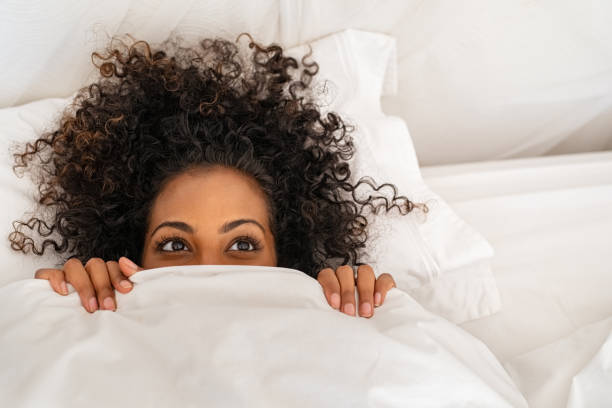 Funny black woman hiding under blanket Funny young woman lying in bed and hiding under sheet while looking up with copy space. Top view of african american girl hiding face under white blanket on bed in the morning. Close up portrait of beautiful woman with curly hair covering face with bed sheet. relaxation lying on back women enjoyment stock pictures, royalty-free photos & images