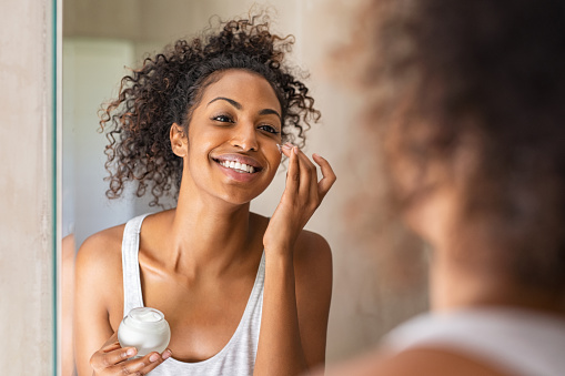 Black woman take care of her beautiful skin. Young african woman applying moisturizer on her face while standing in front of the mirror. Smiling black natural girl holding little jar of skin lotion in bathroom for beauty treatment routine.