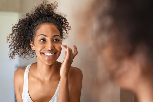 Young black woman applying moisturising cream to her skin while standing in front of the mirror in the bathroom. African american girl applying face cream while smiling. Beauty hydrating moisturizer and skincare routine concept.