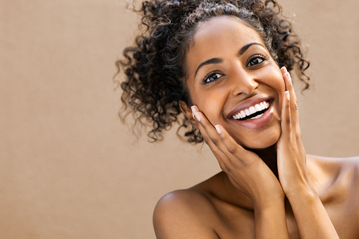 Beautiful young woman smiling after fantastic face treatment . Happy beauty african girl with curly hair and toothy smile excited after spa treatment isolated on background with copy space. Surpise and astonishment beauty concept.