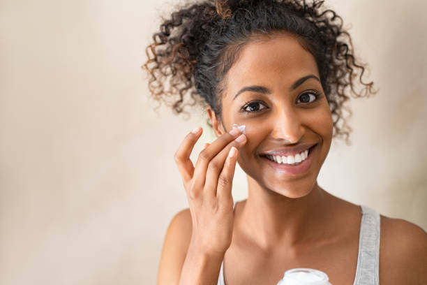 Beautiful girl applying face cream Smiling african girl with curly hair applying facial moisturizer while holding jar and looking at camera. Portrait of young black woman applying cream on her face isolated on beige background. Close up of happy attractive beauty woman caring of her skin standing on light brown wall with copy space. antiaging stock pictures, royalty-free photos & images