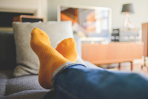 Relaxing at home. Man wearing orange socks has his legs crossed, lying out on the sofa, watching the TV or listening to music on the hi-fi in the background.