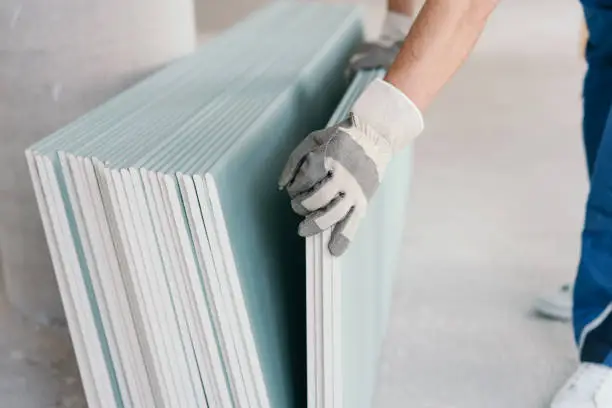 Builder taking a sheet of chip board cladding from a stack against the wall with his gloved hands indoors in a new build home