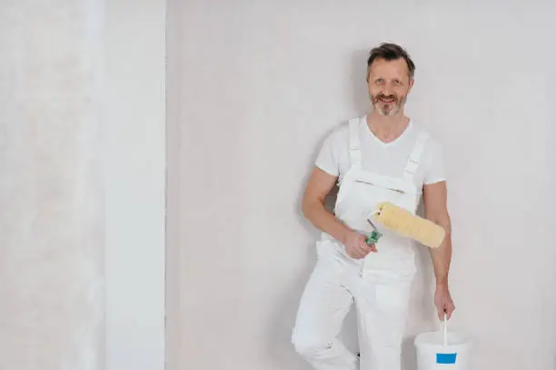 Painter in white dungarees holding a roller and tub of paint as he stands against a white wall smiling at the camera