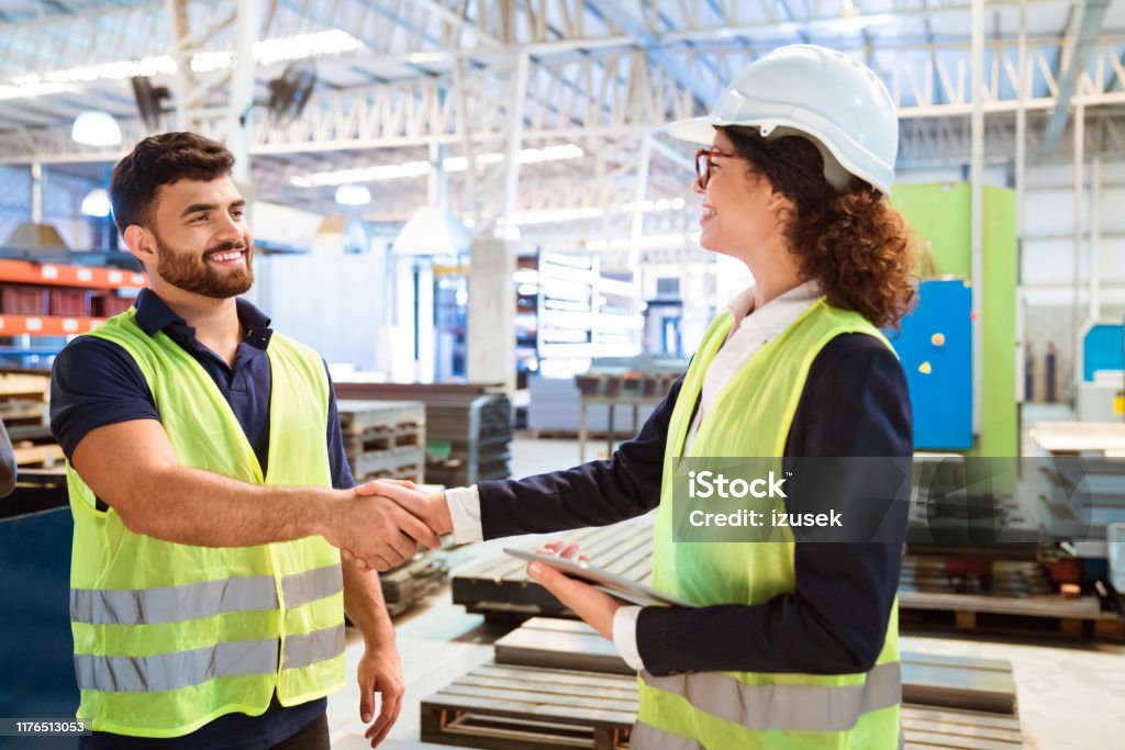 Female manager shaking hand with worker in factory Smiling female manager shaking hand with production worker in factory. Expertise are working in manufacturing company. They are wearing reflective clothing. Factory Stock Photo