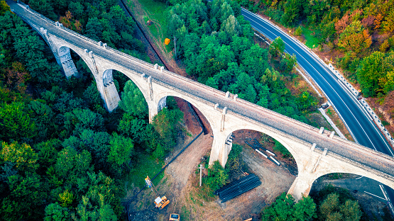 High angle color image taken with drone, depicting a highway with cars and other traffic, flanked by lush forest, and an old abandoned viaduct bridge. Room for copy space.