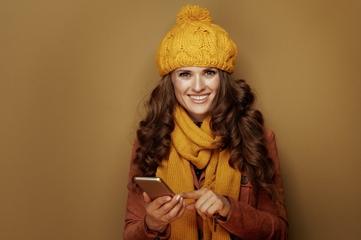 smiling trendy woman in yellow beret and scarf with smartphone hanging in social media and against bronze background.