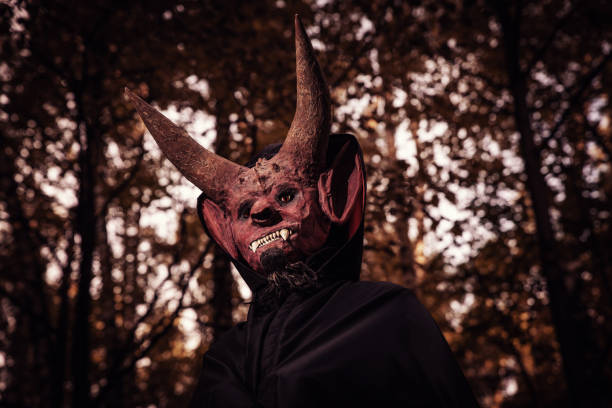 Devil with horns and in a black cloak on a red background. Man in scary mask for Halloween.