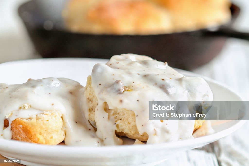 Southern Biscuits and Sausage Gravy American biscuits from scratch covered with thick white sausage gravy. Selective focus with cast iron skillet / pan in the background over a white table. Biscuit - Quick Bread Stock Photo