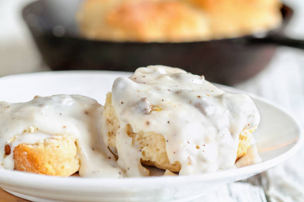 Southern Biscuits and Sausage Gravy American biscuits from scratch covered with thick white sausage gravy. Selective focus with cast iron skillet / pan in the background over a white table. gravy photos stock pictures, royalty-free photos & images