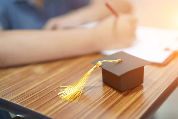 istock Asian children young kid write answer in a quiz examination test study learn with graduation gap hat on wooden table in school : bright genius education concept. 1176545938