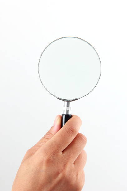 A businessman holds a magnifying glass in front of a long road that disappears into the horizon on a cloud filled day.  The road, as seen through the magnifying glass, is clear and in focus.