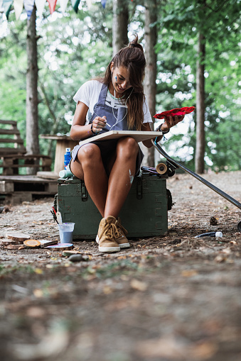 Beautiful and happy young woman enjoying in nature and summer camp. She siitting and painting in the woods.