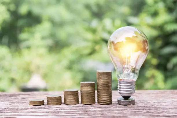 Photo of Energy saving light bulb  stacks of coins on nature background.