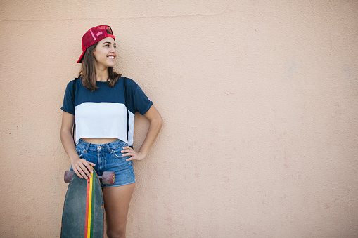 Attractive caucasian teenage girl leaning on the wall and holding long board. Summertime.