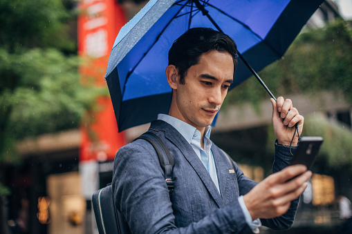 One man, young Taiwanese businessman downtown in Taipei, holding blue umbrella, using smart phone.