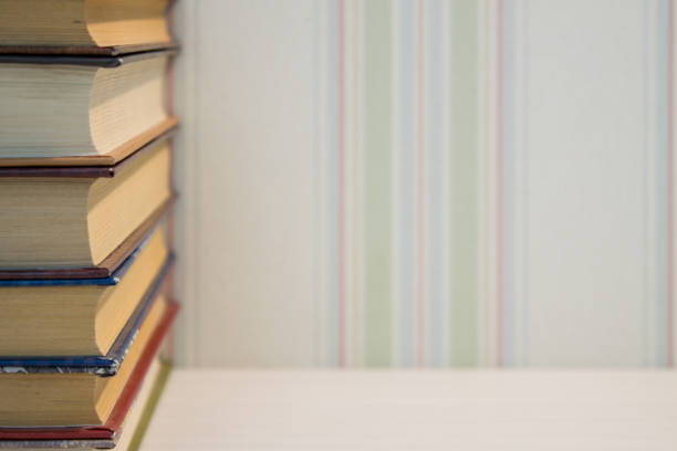 An even stack of old books on the edge of a white wooden table with a free space against the wall with light striped Wallpaper