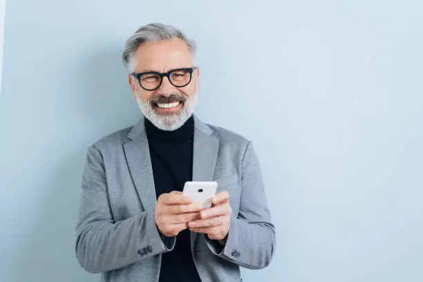 Bearded middle-aged man with grey hair, holding smartphone, looking at camera and laughing. Half-length front portrait against blue wall background with copy space