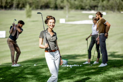 Portrait of an elegant young woman standing with golf putter and friends playing golf on the background