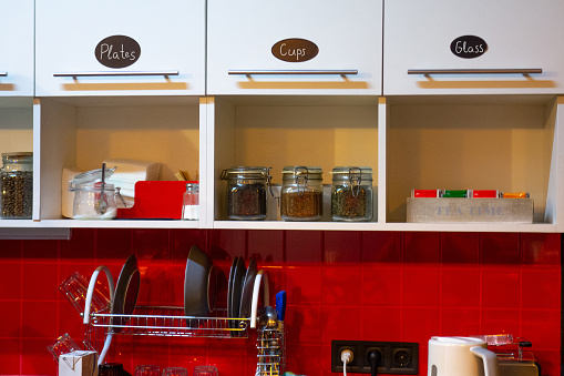 Kitchen interior at home or small office. Red tile wall and white shelf. Cabinet doors with text labels. Glass jars with coffee, tea, sugar. Plates and cups on dish rack. Different cooking utensils