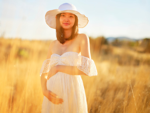 A beautiful pregnant Japanese woman standing in a meadow at dusk with sun flare.
