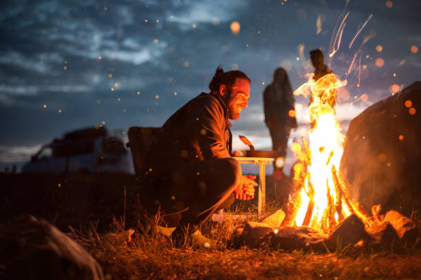 暗闇の中でたき火の隣で微笑む男 - キャンプファイヤー ストックフォトと画像